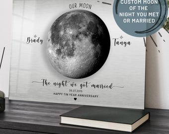 May include: A silver metal print featuring a black and white image of the moon. The text "Our Moon" is above the moon and the text "The night we got married 09.07.2015 Happy Tin Year Anniversary" is below the moon. The text "Brady" is to the left of the moon and the text "Tanya" is to the right of the moon. The text "Custom Moon of the Night You Met or Married" is in a circle in the top right corner of the image.