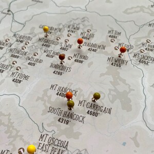 May include: A map of mountains with push pins marking locations. The pins are orange, yellow, and green. The map is printed on a light brown background.