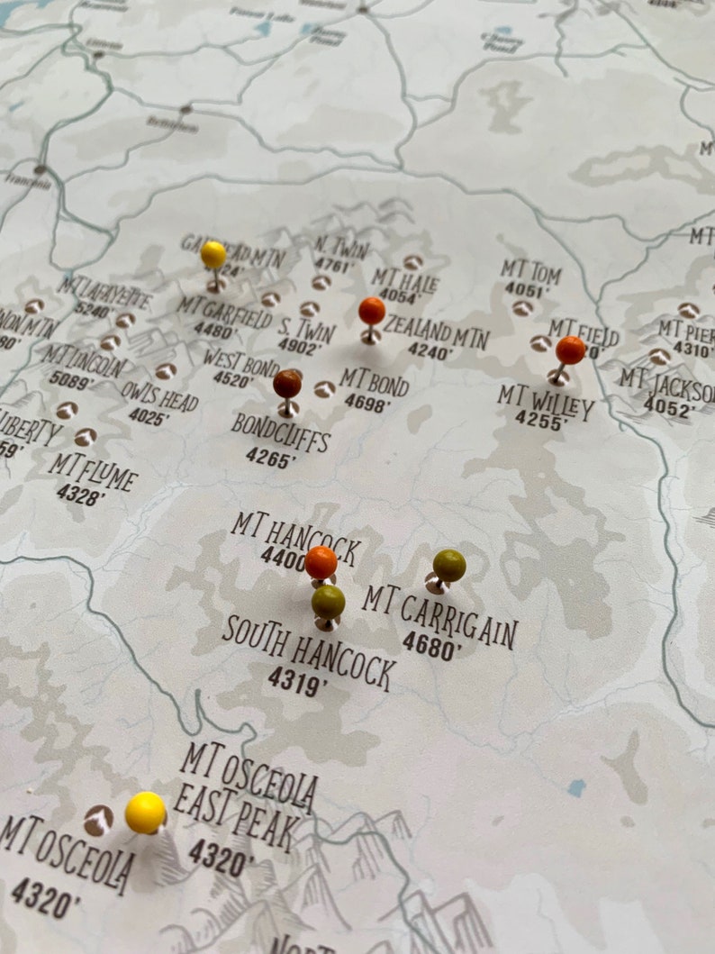 May include: A map of mountains with push pins marking locations. The pins are orange, yellow, and green. The map is printed on a light brown background.