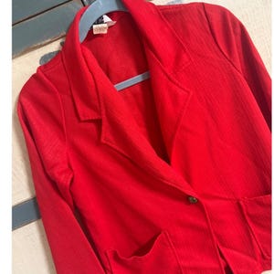 May include: A bright red blazer with a notched lapel, two front pockets, and a single button closure. The blazer is hanging on a blue hanger. The fabric has a subtle ribbed texture.