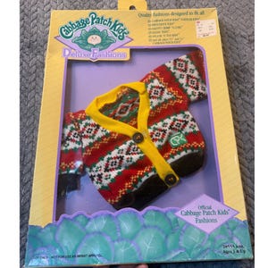 May include: A Cabbage Patch Kids Deluxe Fashions outfit, featuring a red, green, and white patterned sweater with a yellow collar and buttons. The packaging includes the Cabbage Patch Kids logo and text, "Official Cabbage Patch Kids Fashions."