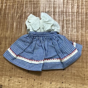 May include: A doll dress featuring a white short-sleeved bodice with a textured fabric and a blue and white gingham skirt. The skirt is accented with a horizontal white ribbon trim and a thin red decorative cord detail along the hemline.