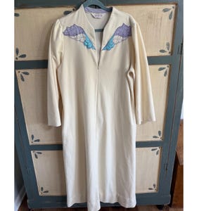 May include: Cream-colored vintage robe with long sleeves and a V-neck. The robe features decorative wing-shaped embroidery in shades of purple and blue on the shoulders. The robe has a full-length zipper closure.