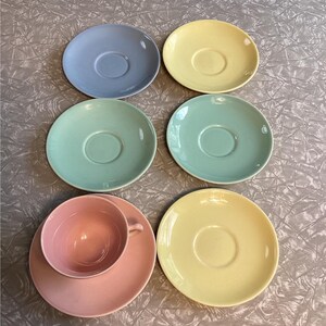 May include: A collection of pastel-colored ceramic saucers and a teacup. The saucers are in shades of blue, yellow, and green. A pink teacup sits on a matching saucer. The items are arranged on a textured, light gray surface.