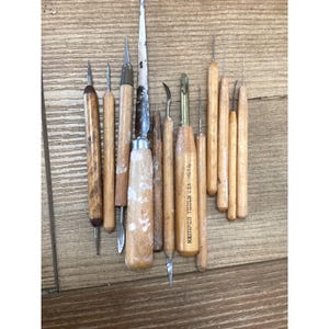 May include: A collection of eleven pottery and sculpting tools with wooden handles of varying shades and textures. The tools feature diverse metal tips, including needles, loops, and blades. One tool is clearly marked with the text Kemper Tools USA H01A.