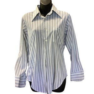 May include: A long-sleeve button-down shirt featuring a white base with vertical stripes composed of small blue geometric patterns. The shirt includes a pointed collar, a single chest pocket on the left side, and wide cuffs with button closures.