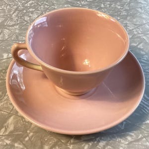 May include: A vintage pink teacup and saucer set. The cup has a rounded shape with a curved handle. The saucer is circular, and the set is a soft, solid pink color. The set is made of ceramic.