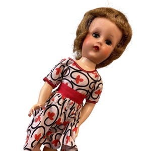 May include: Vintage doll with brown hair and a red and white dress. The dress features a red waist band and a pattern of red flowers and black swirls. The doll has rosy cheeks and red lips.