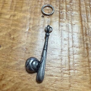 May include: A silver baseball bat and ball charm with a small ring above it. The charm is detailed with stitching on the ball and a textured handle on the bat. The charm is likely for a necklace or bracelet.
