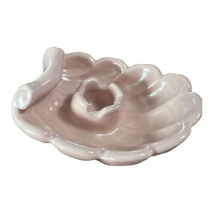 May include: A light pink ceramic seashell-shaped dish with a small, round holder and a curved handle. The dish has a scalloped edge and a glossy finish, perfect for holding small items.