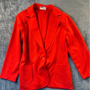 May include: Bright red blazer with a notched collar, two front pockets, and a single-button closure. The blazer has long sleeves and a tailored fit. The fabric appears to be a smooth, possibly knit material. The blazer is a solid color.
