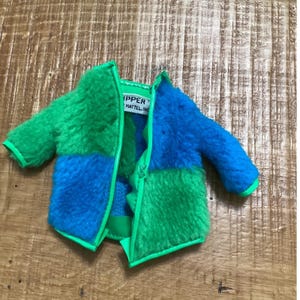 May include: A doll-sized jacket featuring a color-blocked pattern of bright green and blue fuzzy faux fur. The jacket has a neon green trim along the edges and a white interior tag that reads "SKIPPER" and "MATTEL, INC." in black text.
