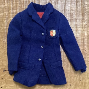 May include: A dark blue blazer with a notched lapel, three silver-toned buttons down the front, and two flap pockets. The jacket features a small shield-shaped crest patch on the left chest and a red interior lining visible at the collar.