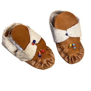 May include: A pair of baby moccasins. The shoes are made of brown leather with a white suede cuff and are decorated with colorful beads. The moccasins are designed with a soft sole and are likely for indoor use.