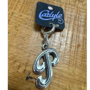May include: Silver-toned metal keychain featuring the letter "P" in a decorative font. The keychain is attached to a black tag with the name "Carlyle" in a script font. The background is a wooden surface.