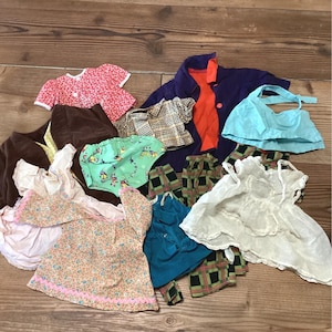May include: A collection of doll clothing including a red floral top, a purple jacket with orange lining, a light blue halter top, a brown skirt, a green patterned pair of shorts, a white dress, a tan floral top, and plaid trousers in various colors.