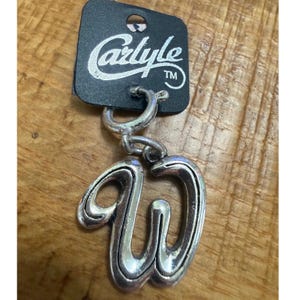 May include: Silver-toned metal earring with a cursive "W" charm, hanging from a small hoop. The earring is attached to a black tag with the name "Carlyle" in white script. The background is a wooden surface.