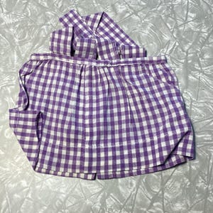 May include: A purple and white gingham apron with straps. The apron is made of a lightweight fabric and features a classic checkered pattern. The apron is designed to be worn while cooking or doing other household chores.