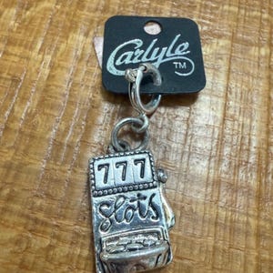 May include: Silver-toned charm featuring a slot machine design with the numbers "777" and the word "Slots". The charm is attached to a black tag with the name "Carlyle" printed on it. The charm is suspended from a silver hoop.