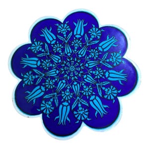 May include: A round, cobalt blue ceramic coaster with a floral design. The coaster features a central floral pattern surrounded by stylized turquoise tulip and leaf motifs. The edges are scalloped and outlined in white.