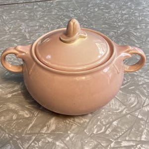 May include: A vintage pink ceramic sugar bowl with a lid and two handles. The lid has a decorative finial. The bowl is round and has a glossy finish. The handles are curved and have a decorative design.