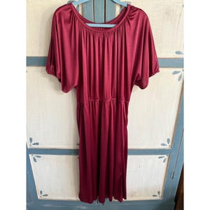 May include: A burgundy-colored dress with short sleeves and a gathered waist. The dress has a round neckline and a flowing skirt. The fabric appears to be a soft, silky material. The dress is hanging on a blue hanger.