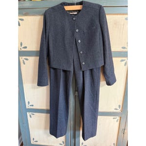 May include: A charcoal gray two-piece suit. The jacket has a round neckline, long sleeves, and a button-up front. The matching pants are straight-legged. The suit is hanging on a wooden hanger against a light blue cabinet.