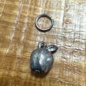 May include: A silver-colored apple charm with a small leaf and a loop for attaching to a necklace or bracelet. A silver-colored ring is above the charm. The charm and ring are on a wooden surface.