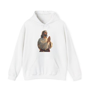 May include: White hoodie with a graphic of a person with long hair and a beard, hands clasped in prayer. The graphic is in brown, beige, and white. The sweatshirt has a front pocket and a drawstring hood.