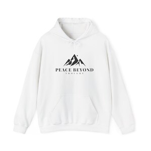 May include: White hoodie with a front pocket and drawstring. The front displays a black mountain graphic and the text "PEACE BEYOND THOUGHT". A casual, comfortable garment for everyday wear.