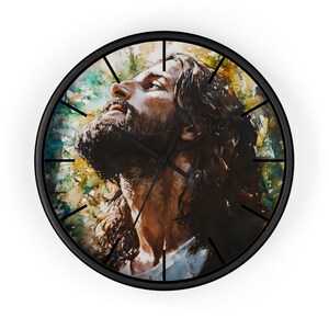 May include: Round wall clock with a black frame featuring a watercolor portrait of a man with long hair and a beard looking upwards. The background has green, yellow, and blue hues. Black clock hands indicate the time.