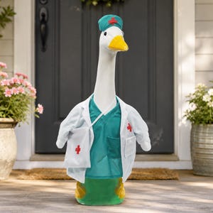 May include: A white goose figurine dressed as a medical professional, wearing a teal scrub top, a white lab coat with red cross emblems, and a teal surgical cap with a red cross. The goose has a yellow beak and black eyes, standing upright.