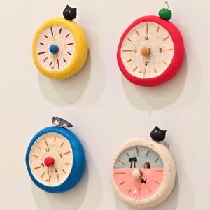 May include: Four colorful, whimsical clock magnets. Each clock has a different color scheme: yellow, red, blue, and off-white with pink. Each clock has a small decorative element on top, such as a black cat or a green berry.