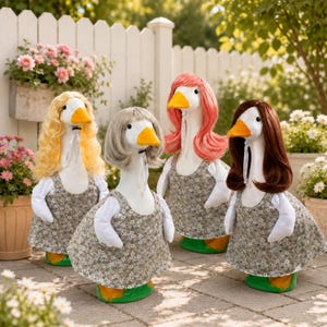 May include: Four white goose figurines standing on a paved surface, each wearing a gray floral print dress with white sleeves. The geese have different colored synthetic hair wigs: one blonde, one gray, one pink, and one brown, all secured with ribbons.
