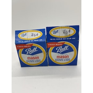 May include: Two blue boxes of Ball mason jar dome lids. Each box has a yellow and white circular logo with the word "Ball" in cursive. The text "12 Regular Mason Dome Lids" is below the logo. The boxes also have the text "Vacu-Seal".