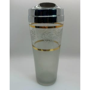 Può includere: Un vintage cocktail shaker con top e base argentati, e un corpo in vetro trasparente. Il vetro è decorato con una fascia dorata e un motivo floreale. Lo shaker misura circa 25 cm di altezza.