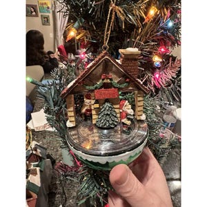 May include: A Christmas ornament shaped like a cabin with a chimney, a small Christmas tree, and a Santa Claus figure. The ornament is round with a green and white base and is hanging on a Christmas tree.