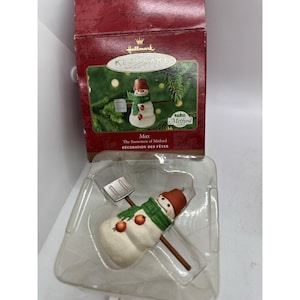 May include: A Hallmark Keepsake Christmas ornament featuring a snowman named Max. The snowman has a brown hat, green scarf, and orange accents. The ornament comes in a red box with the text "Max The Snowmen of Mitford".