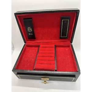 May include: Open black jewelry box with a red velvet interior. The box has a gold-colored clasp and trim. The interior features ring slots and compartments. The lid has a black panel with gold lettering and a crest.