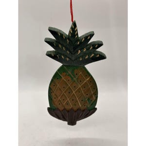 Puede incluir: Un adorno de madera tallada con forma de piña que presenta una parte superior de hojas verde oscuro con detalles dorados, un cuerpo texturizado en tonos marrón y verde con patrón de rombos y una base marrón oscuro, colgado de un hilo rojo.