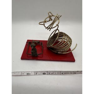 May include: A whimsical sculpture of a mouse and a mousetrap. The mouse is made of brass wire and sits on a red rectangular base. The mousetrap is also brass and is positioned near the mouse. The sculpture is approximately 5 inches long.