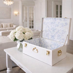 May include: A white rectangular storage chest with gold-toned metal latches and handles. The interior lid and walls feature a light blue and white floral pattern. The text The Monroes is printed in white script on the inside of the open lid.