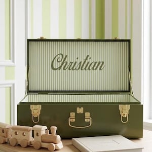 May include: An open, olive green trunk with gold hardware, the interior lined with green and white stripes and the name "Christian" in script. A wooden toy train and a book sit on a light-colored table.