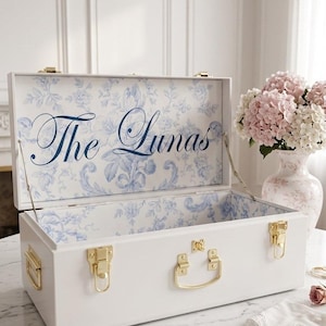 May include: White wooden chest with gold hardware, open to reveal a blue floral patterned interior. The text "The Lunas" is displayed inside the lid. A vase of pink flowers sits nearby on a marble table.