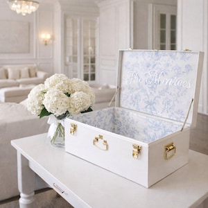 May include: White decorative trunk with gold-colored hardware, open to reveal a blue and white floral patterned interior. The inside lid features the embroidered name "The Monroes". A bouquet of white flowers sits nearby on a white table.