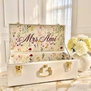 May include: A white rectangular box with gold-colored hardware, including a handle and clasps. The interior and the inside of the lid are lined with a floral pattern. The lid is open, revealing the name "Mrs. Ami" in burgundy script.