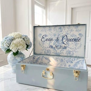 May include: A light blue rectangular storage chest with gold-toned metal latches and a handle. The interior lid features a blue and white floral pattern with the text Evan & Queenie and the date 06.20.2026 printed in a dark blue script font.