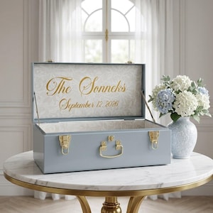 May include: A light blue rectangular metal storage chest with gold-colored latches and a central handle. The interior lid features a white floral pattern with the gold text The Sonneks and the date September 12, 2026, printed in a script font.