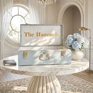 May include: A light blue rectangular storage trunk with gold-toned metal latches and a central handle. The interior lid features a white floral line art pattern and the gold text The Hammels printed in the center of the open lid interior.