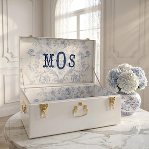 May include: A white rectangular storage trunk with gold-toned metal latches and a handle. The interior is lined with a blue and white floral pattern, featuring the monogram letters MOS in a dark blue serif font centered on the inside lid.